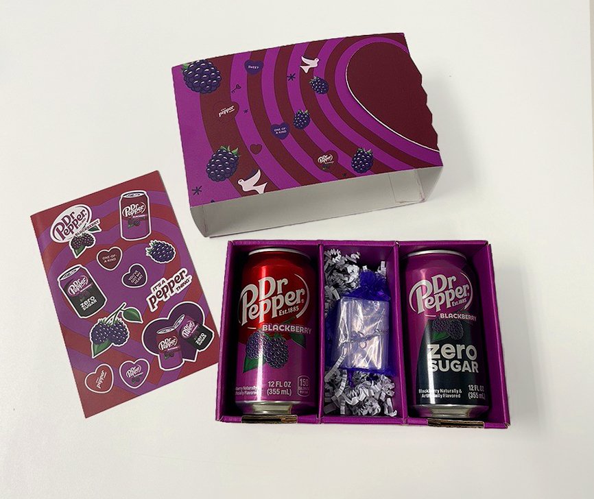 Overhead view of the Dr Pepper Blackberry influencer kit with original and zero sugar cans, a gift pouch, heart patterned lid, and sticker sheet